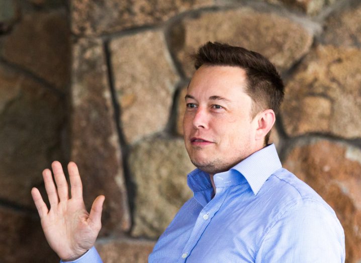 Elon Musk in a blue shirt waving with his right hand, with a brown wall behind him.