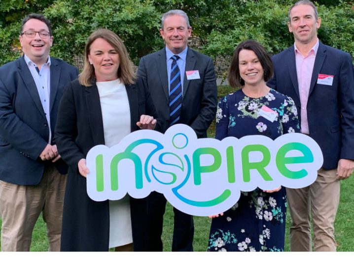 Five people, two women and three men standing in a row outside in front of green trees holding a banner that says Inspire.