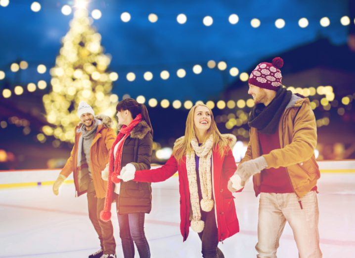 A group of people skating on ice while holding hands, with lights and a Christmas tree in the background.