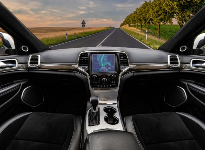 Interior of a self driving car with no steering wheel or driver, going down a road with a field, trees and a cloudy sky in the background.