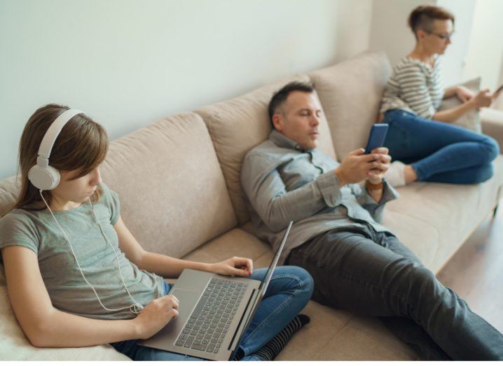 Three people lounging on a sofa in a living room setting. They are looking at their internet devices and a young girl is wearing headphones.