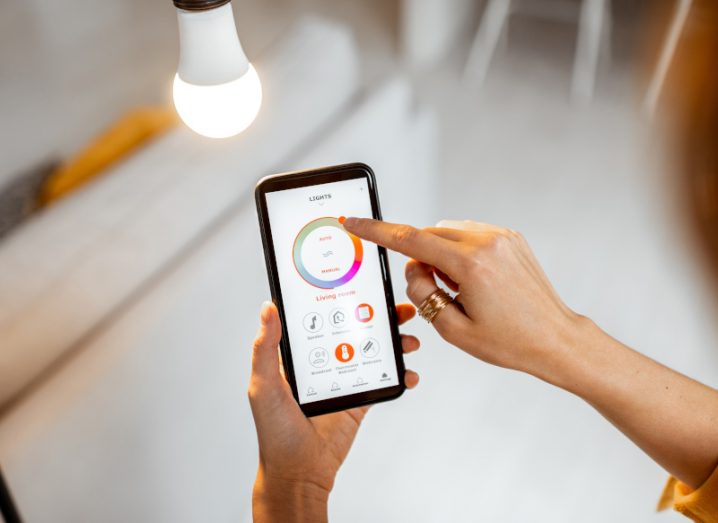 Close-up of a smartphone as a user adjusts the settings of a smart lightbulb via an app. The bulb is lighting the room in the background.