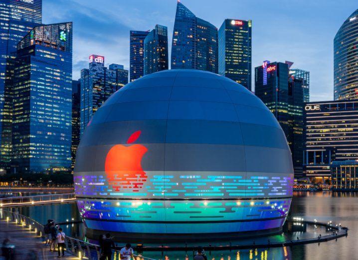 An Apple company logo on the side of a spherical building, with blue lights surrounding the bottom of it. There is a city in the background.