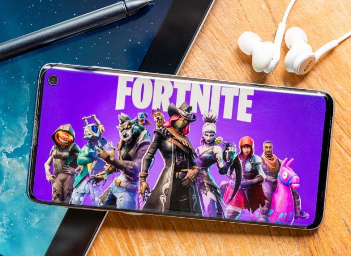 Fortnite logo on a smart phone screen, resting on a wooden table next to a pair of earphones. The game is created by Epic Games.