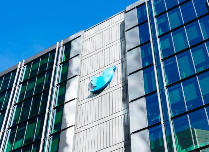 The Twitter logo on the front of a grey building, with windows on each side and a blue sky above the building.