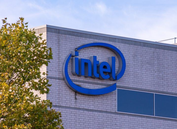 Intel company logo on the side of a grey building, with a tree visible to the left and a blue sky in the background above.