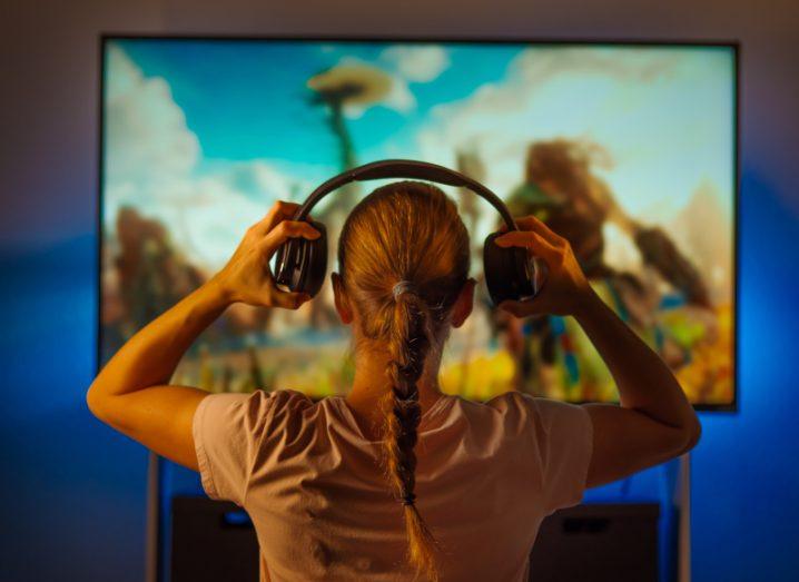 A girl putting a pair of headphones on her head while looking at a TV in the background.