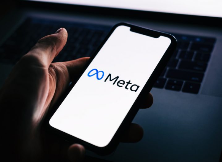 Meta logo on a smartphone in a person's hand. There is a laptop in the background, with light coming from the screen.