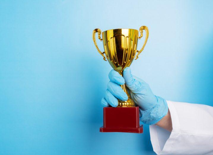 A gold trophy being held in the hand of a person wearing a white medical coat and a blue glove, in a bright blue background.