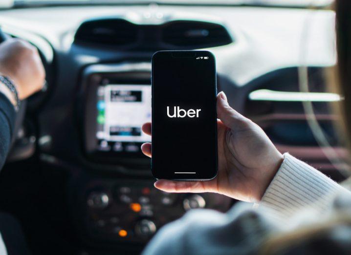 A smartphone with the Uber logo on the screen, being held in a person's hand. The phone is inside a car, with a person's hand visible on the steering wheel.