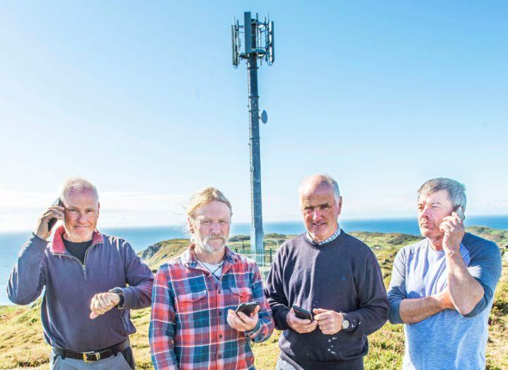 Four men standing in front of a comms tower on Cape Clear island using their mobile phones. The sea is visible behind them.