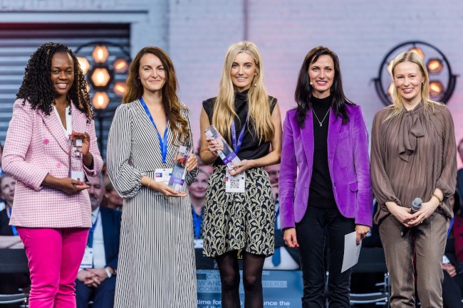 Five women stand on a stage, three of whom are holding awards. Niamh Donnelly, in the centre next to EU commissioner Mariya Gabriel, is smiling.