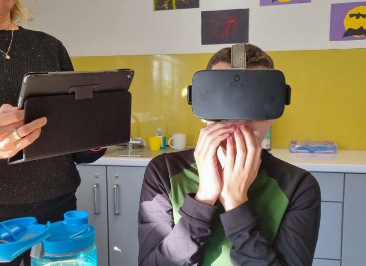 A child wearing a VR headset in a classroom, with an adult standing nearby holding a digital tablet.