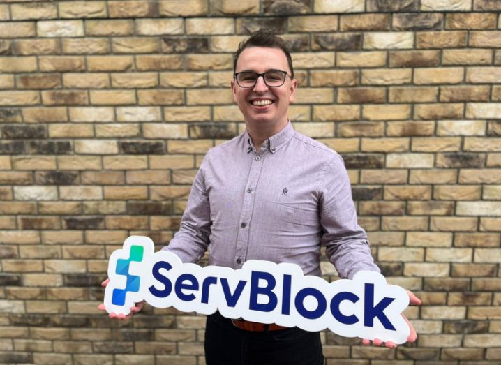 John Ward standing in front of a brick wall and holding a ServBlock sign in his hands.