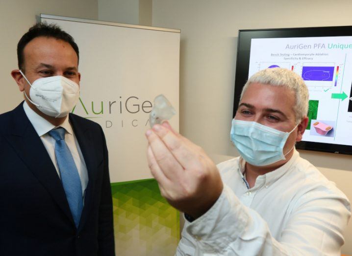 Two men looking at a small medical device, being held out by the man on the right. They are both wearing face masks and the AuriGen Medical logo is on a small sign in the background.
