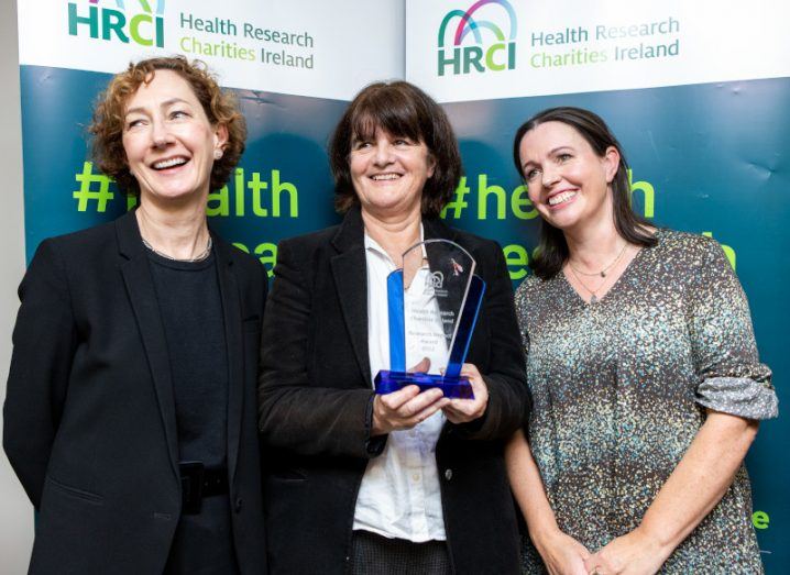 Three women stand together while the one in the centre holds an award.
