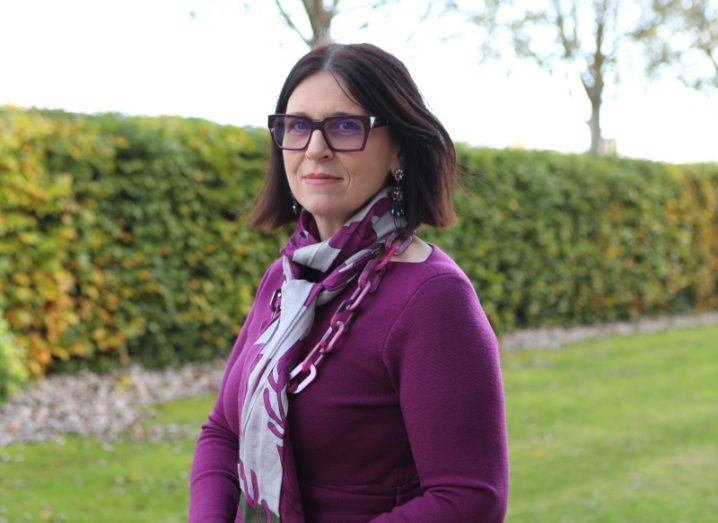 A woman with dark hair and glasses smiles at the camera while standing outdoors with hedges behind her.