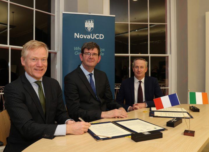 Three men seated at a table signing a document. The flags of France and Ireland can be seen on the table.