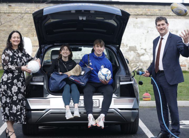 Two people sitting in the boot of a car holding sports gear while two other stand on either side of the boot.