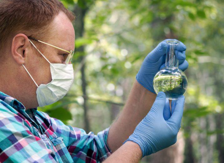 A man wearing a face mask and holding a glass bottle containing a clear liquid. The man is wearing blue gloves and is in the woods, with trees in the background.