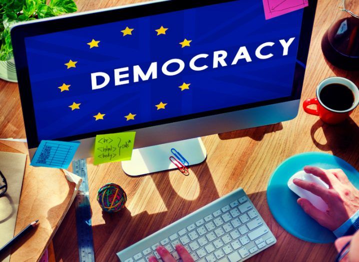 People's hands visible in front of a computer monitor with the EU flag and the word democracy written on the screen.