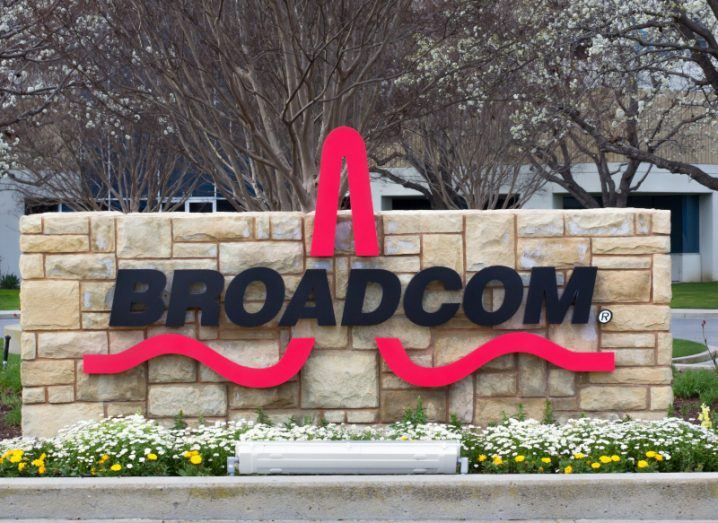 The Broadcom logo in black letters, placed in front of tan bricks, with flowers in front and bare trees in the background.