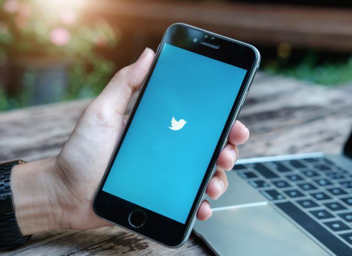 Twitter logo on a smartphone screen, being held in a person's hand. There is a laptop resting on a table behind the phone.
