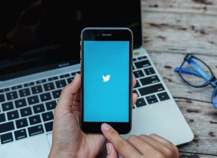 Twitter logo in a blue background on a smartphone screen, held in a person's hand. A laptop is behind the phone, resting on a wooden table next to a pair of glasses.