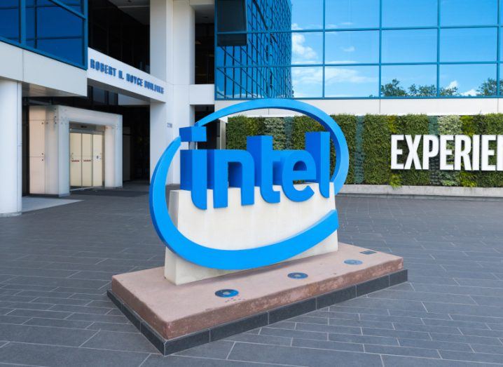 The Intel logo on a sign in front of a building entrance. There are green hedges in the background with a sign saying "Experience" on them.