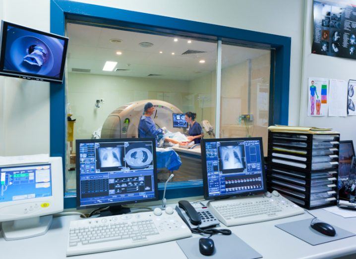 A medical room with computer screens showing various images from an MRI machine. The machine can be seen in the background through a window, with a patient laying down and two doctors standing alongside.
