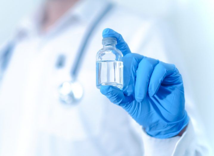 A person in a doctor's outfit holding a small vial of liquid, wearing blue surgical gloves.