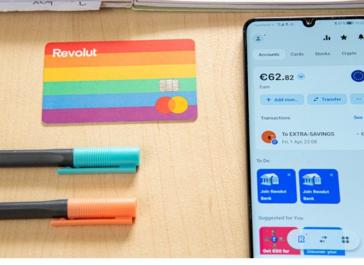 Revolut app open on a phone with a rainbow-coloured Revolut card beside it and two pens.