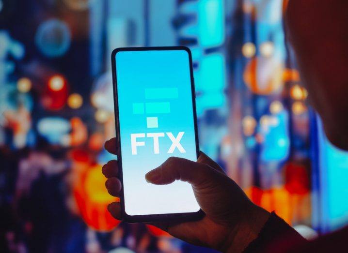 The FTX logo on a smartphone screen held in a person's hand with a city in the background.