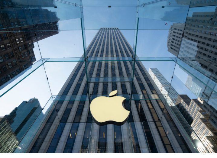 View from the ground of a large skyscraper with the Apple logo on the building against a blue sky background.