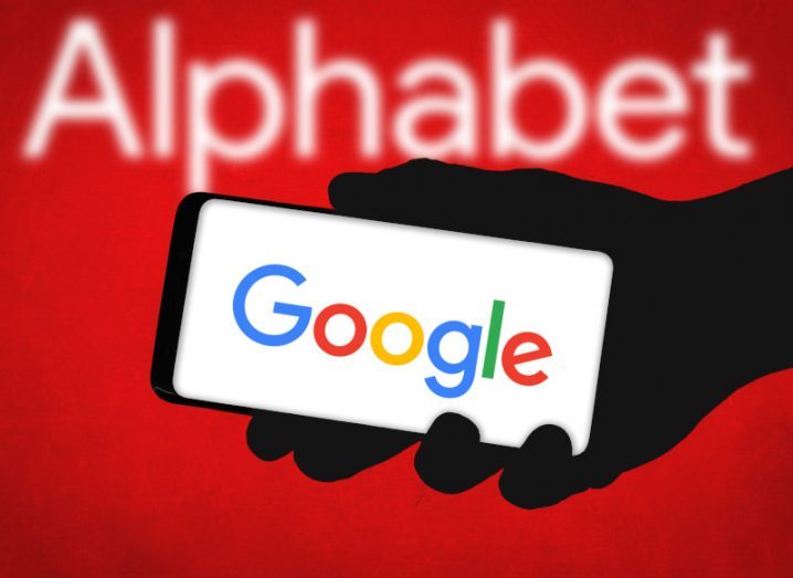 The word Alphabet written in white letters on a red background, with a person's hand holding a phone that has Google's logo on the screen reaching out in front.