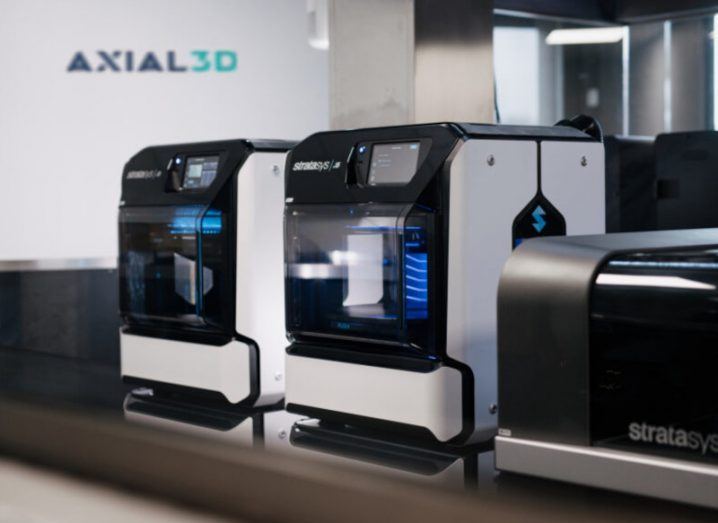 Two 3D printing devices in an office, with the Axial3D logo on a wall in the background.