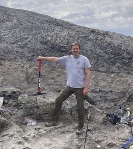 Ben Kligman, paleontologist, poses for a picture at a dig sit with a shovel in his hand, wearing a grey t-shirt and dark trousers. There are hills and a cloudy sky in the background.