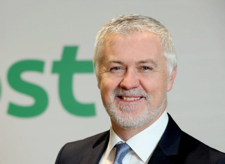A man in a dark suit smiling at the camera. Behind him, a wall has a green S and T on it, which is the end of the An Post logo.