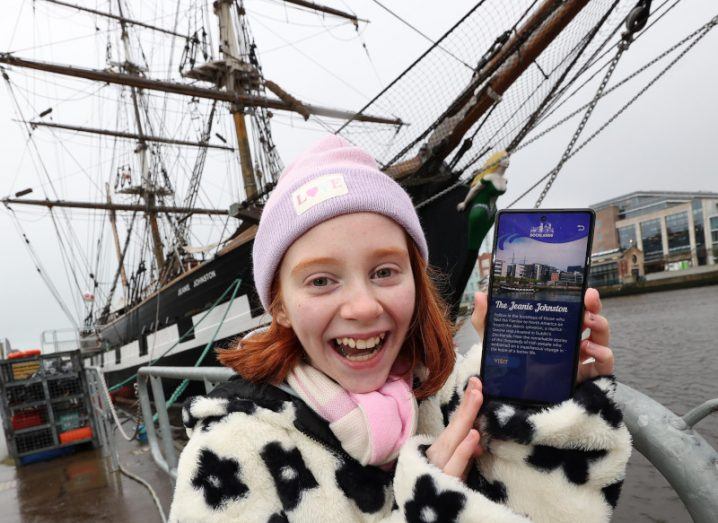 A little girl standing in front of the Jeanie Johnston ship holding a smartphone with the Dublin Docklands AR app on it.