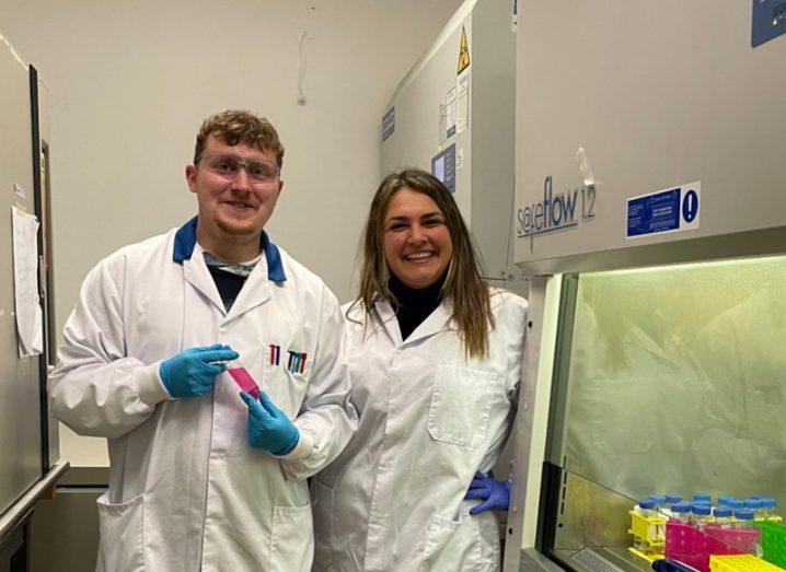 A man and a woman in white lab coats, standing together in a small lab room.