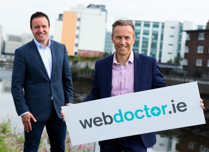 Two men in suits standing in front of a river, with the man on the right holding a sign for WebDoctor.ie.