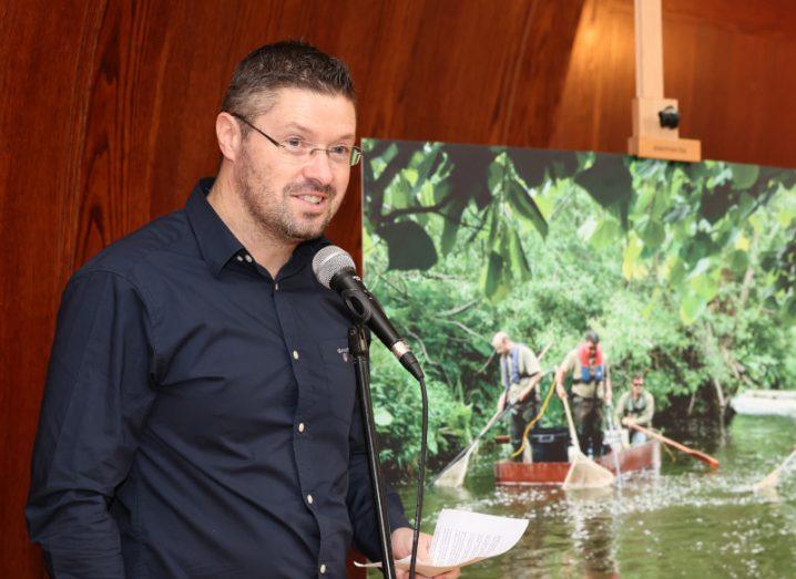 A man stands at a microphone. Beside him is an easel holding an image of people in boats along the River Rye.