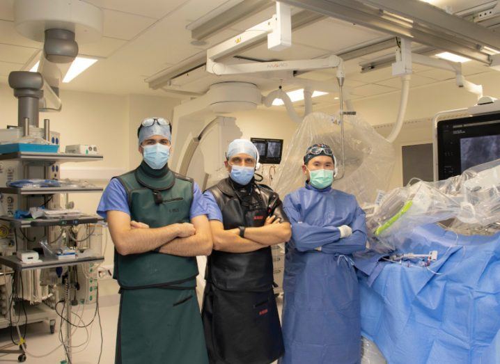 Three surgeons wearing masks, standing together in a surgery room. There is equipment and a blue cloth behind them.