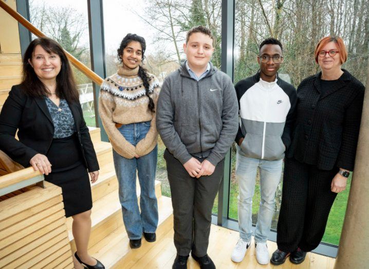 The three teen winners of TECS with a professor on either side of them standing beside a stairway with a glass window showing green trees outside.
