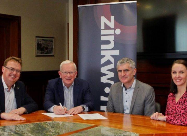 Three men and one woman sit around a circular table. One man in the middle is signing a piece of paper. The Zinkworks banner is behind them.