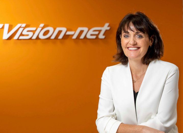 Woman wearing a white blazer sits by a table and smiles. An orange wall behind her has 'Vision-net' written on it.