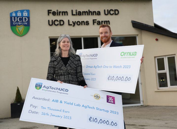 Fiona Kelleher and Gary Gallagher smile and hold up large cheques with the AgTech UCD logo. They are standing in front of the UCD Lyons Farm building.