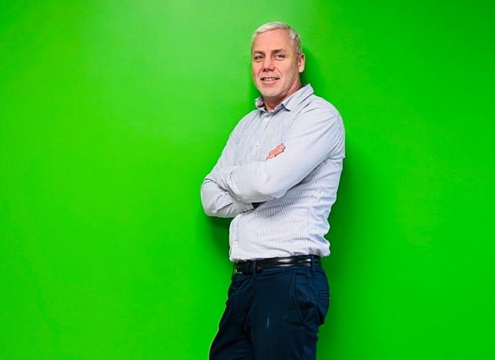 A man wearing a white shirt and black trousers stands against a bright green wall with his arms folded.