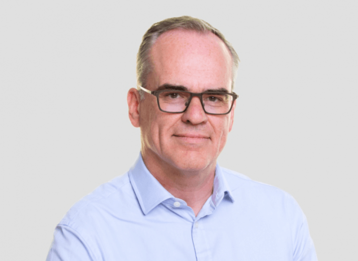 Headshot of Kyndryl's John Chambers. He is wearing a blue shirt and is pictured against a grey background.