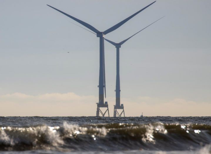 Two offshore wind turbines off the coast of Aberdeen. There are waves in the foreground and a clear, calm sky in the background.